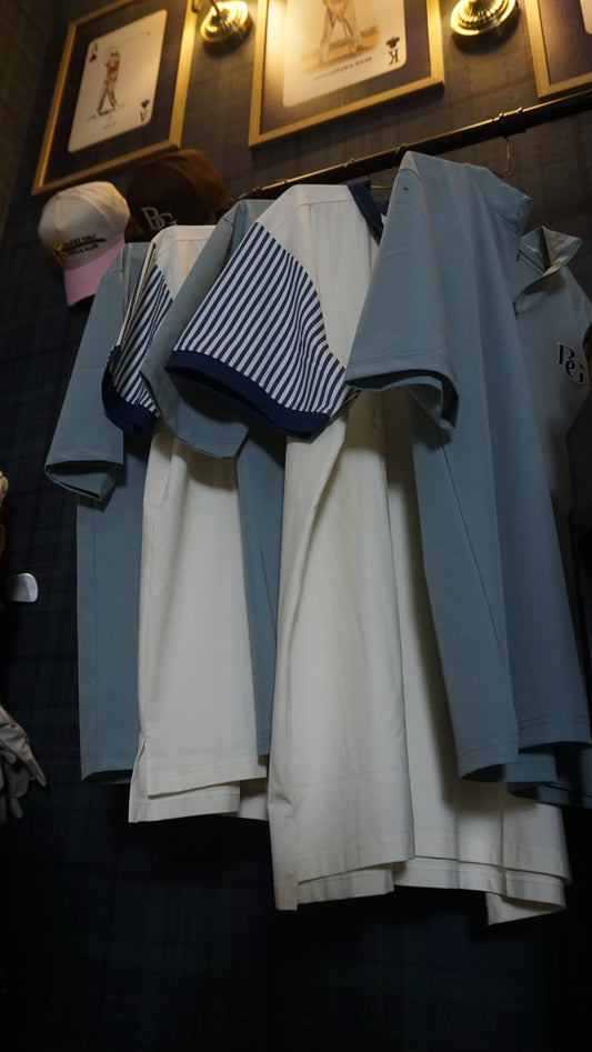 A set of golf apparel tops hanging on a rack in a dimly lit room. The set includes light blue short-sleeve 1/4 zip windbreaker, and blue-and-white striped short-sleeve polo.