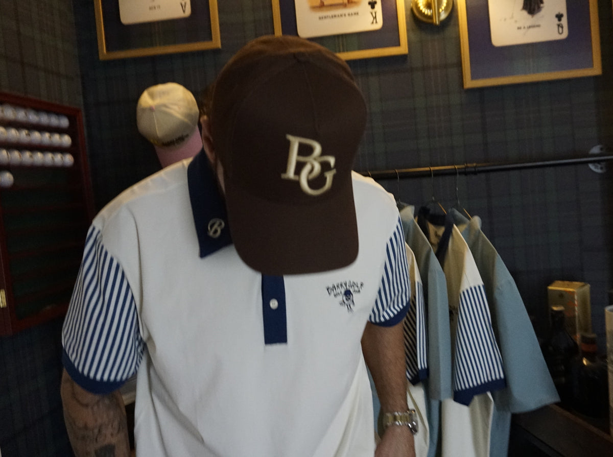 A man wearing a brown baseball cap with a gold 'BG' logo and a white polo shirt with blue striped sleeves, standing in a retail store.