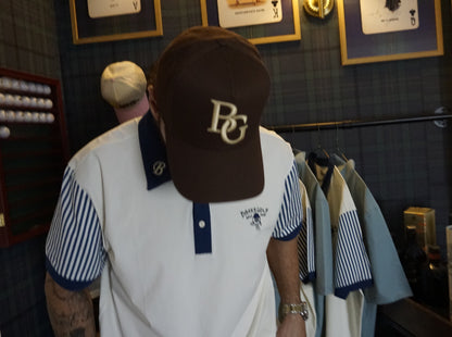 A man wearing a brown baseball cap with a gold 'BG' logo and a white polo shirt with blue striped sleeves, standing in a retail store.
