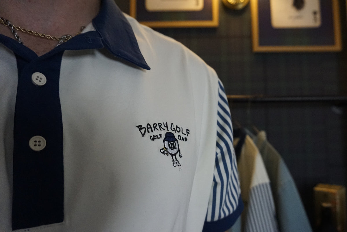 A close-up of a person wearing a white polo shirt with a navy blue collar and placket, featuring a navy blue embroidered logo that reads 'BARRY GOLF, GOLF CLUB' with a small golf ball character. The right sleeve has navy blue and white vertical stripes.