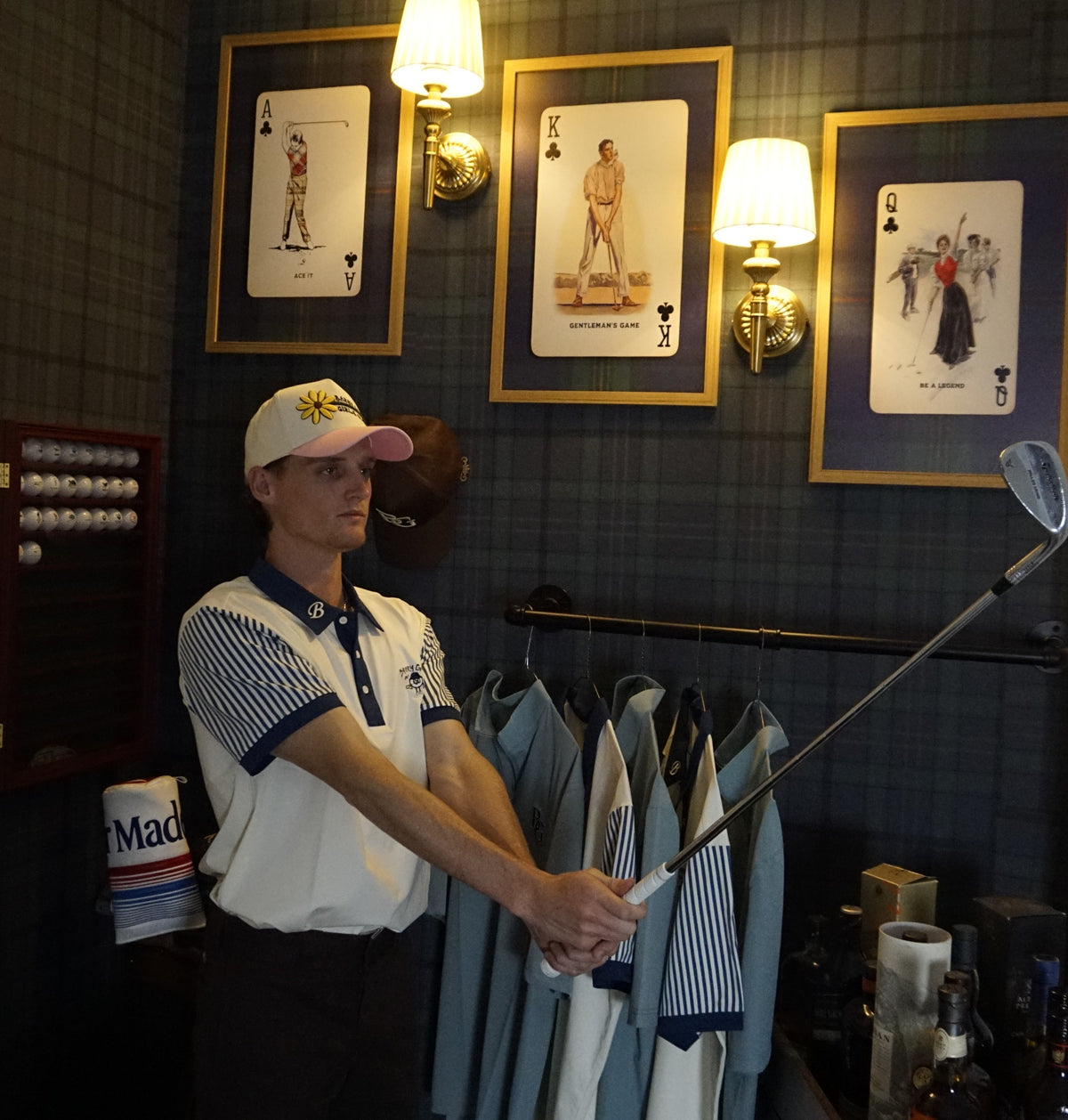 A man in a golf-themed room wearing a white and navy striped polo shirt and a cap, holding a golf club. Golf shirts hang on a rack against a dark plaid wall with framed playing-card style art and wall sconces.